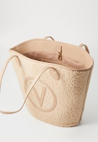 Ljusbeige tote-väska med plyschshearling exteriör och släta läderdetaljer. Har en logotyp, två handtag och en innerficka.