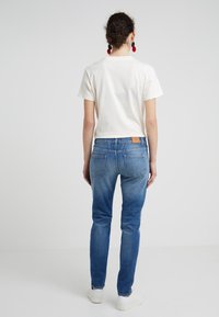 Bílé tričko s krátkým rukávem, modré džíny s lehkým vyblednutím a bílé tenisky. Viditelné zadní kapsy, šité detaily a kožená nášivka.