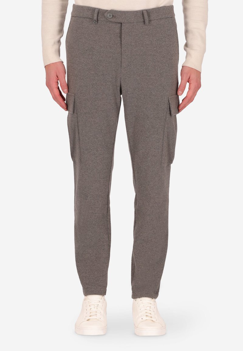 Uomo che indossa pantaloni cargo grigi con tasche laterali e sneakers bianche, in piedi su uno sfondo bianco.