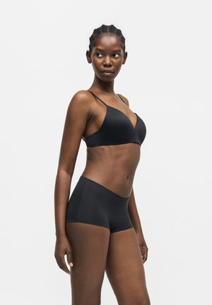 Femeie slabă cu părul împletit, purtând un sutien negru și chiloți boyshort cu talie înaltă asortați, stând în picioare și uitându-se spre stânga.