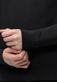 Mains à la peau claire ajustant le poignet côtelé d’un pull noir à manches longues sur fond noir.