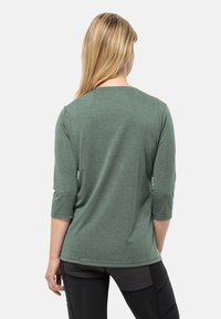 Maglietta verde a maniche 3/4 realizzata in tessuto morbido e testurizzato, con scollatura arrotondata e vestibilità ampia, abbinata a leggings sportivi neri.