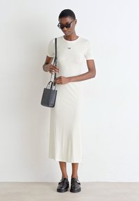 Cream-colored fitted dress with short sleeves and a round neckline, paired with a black handbag and chunky black shoes.