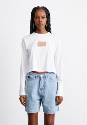 Chemise blanche à manches longues raccourcies avec un logo "SANTA CRUZ", associée à un short en denim bleu clair à coupe classique et fermeture à bouton.