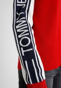 Czerwony sweter z białymi i granatowymi paskami na rękawie oraz wplecionym logo "TOMMY". Miękki materiał dzianinowy z dopasowanym krojem.