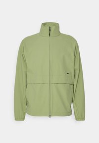 Chaqueta Nike de color verde claro con un alto cuello, cremallera frontal, mangas raglán y una costura horizontal en el pecho. Textura suave, mínima marca.