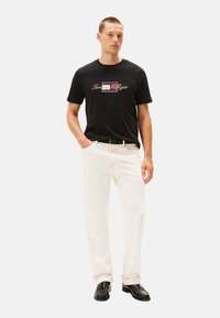 Camisa de algodón negra con un estampado de logo en rojo, blanco y azul; combinada con jeans de mezclilla blancos y zapatos de cuero negros. Corte casual.