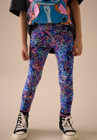 Leggings colorés et extensibles en bleu avec un motif éclatant de taches roses, oranges et vertes. Associés à des baskets montantes noires.
