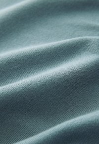 Weicher, strukturierter Stoff in petrol mit dezentem Rippenmuster, der sanfte Wellen und Falten zeigt und ein glattes, leichtes Material betont.