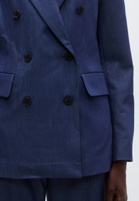 Dvigubai sagaunamas tamsiai mėlynas švarkas su smulkia tekstūra, turintis šešis juodus mygtukus ir dvi priekines kišenes. Siūtas modelis su struktūriška forma.