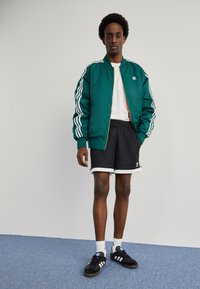 Groene gewatteerde bomberjack met witte strepen, zwarte mesh korte broek met witte accenten, witte t-shirt en zwarte sneakers met blauwe logo.