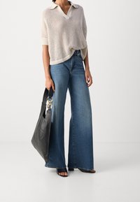 Lys beige strikbluse med korte ærmer og V-hals, parret med viddejeans i blå denim. Holder en sort lædertaske med guldfarvede detaljer.