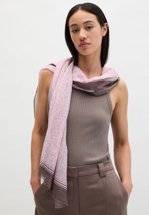 Jonge vrouw draagt een mouwloos geribd topje en een bruine broek, met een roze en bruine sjaal met patroon over haar schouders gedrapeerd.