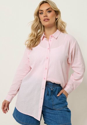 Kvinde med langt blondt hår iført en lyserød og hvid stribet skjorte med knapper og blå denimshorts, poserer med den ene hånd i lommen.