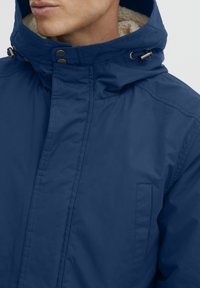 Jūras zila jaka ar mākslīgās kažokādas oderi kapucei, priekšā ar rāvējslēdzēju, pogu aizdari un sānu kabatām. Audumam ir gluda tekstūra.