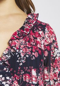 Blus med blommönster som har en volangkrage, i nyanser av marinblå, rosa, röd och vit. Lättviktigt tyg med en genomskinlig struktur.