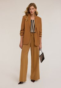 Blazer marrone su misura sopra una maglietta a righe, abbinato a pantaloni beige a gamba larga. Completano il look una piccola borsa nera e décolleté a punta.