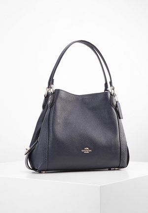 Sac à main Coach en cuir bleu foncé avec deux poignées, logo doré sur le devant, et une petite étiquette attachée, présenté sur une surface blanche.