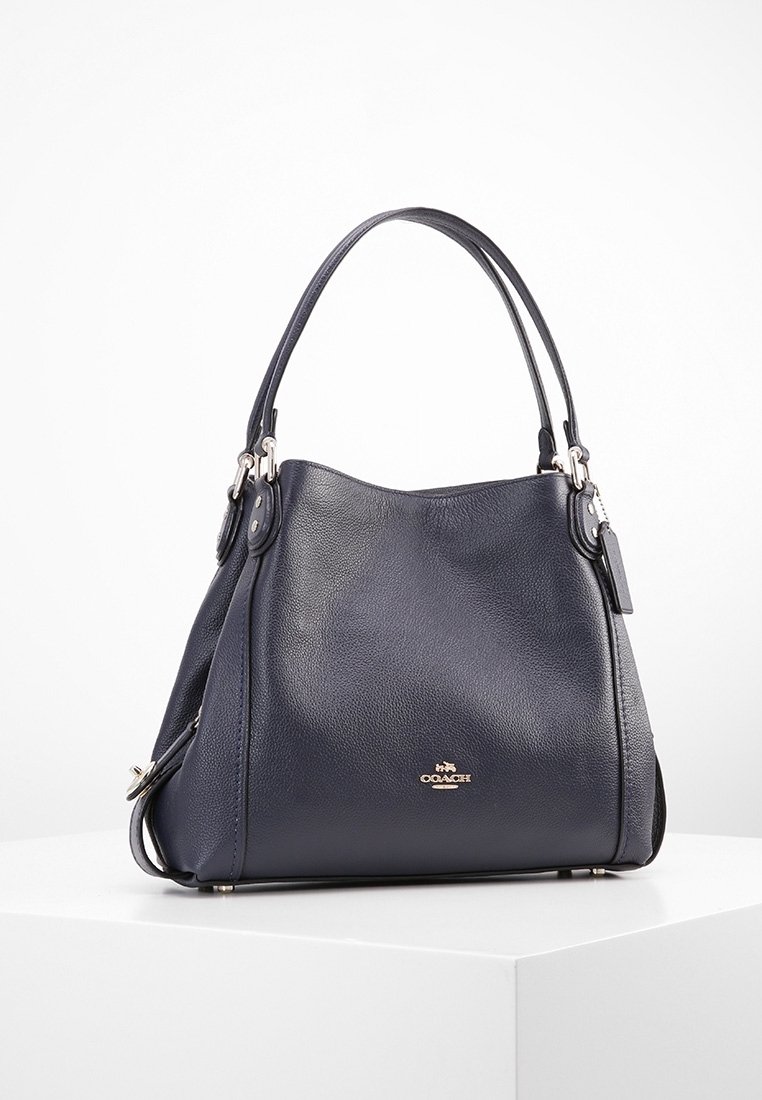 Sac à main Coach en cuir bleu foncé avec deux poignées, logo doré sur le devant, et une petite étiquette attachée, présenté sur une surface blanche.