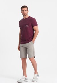 Camiseta color burdeos con un bolsillo frontal, combinada con pantalones cortos grises claros. Los pantalones cortos tienen un dobladillo curvado. Zapatillas blancas con detalles grises.