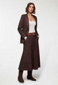 Blazer in suede marrone sopra una maglietta bianca, abbinato a una lunga gonna marrone. Include una cintura marrone e stivali alla caviglia marroni con cuciture decorative.