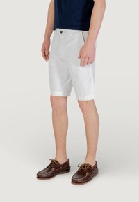 Shorts en coton blancs avec une texture lisse, longueur au-dessus du genou, présentant une fermeture à bouton unique et des ourlets retroussés. Assortis à des chaussures en cuir marron.