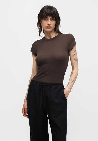 Γυναίκα με σκούρα μαλλιά που φοράει ένα εφαρμοστό καφέ κοντομάνικο τοπ και φαρδιά μαύρα παντελόνια, δείχνοντας τατουάζ στα χέρια της, ποζάρει με το ένα χέρι στην τσέπη.