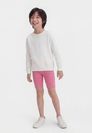 Niño sonriente con una camiseta blanca de manga larga, pantalones cortos rosas con un pequeño logo blanco y zapatillas blancas, de pie frente a un fondo liso.