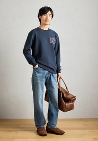 Sweatshirt bleu foncé avec motif, jeans bleu clair, chaussures en daim marron et sac en cuir marron. Design simple et coupe décontractée.