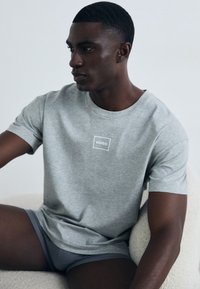 Grå bomulls T-skjorte med en hvit rektangulær logo som står "HUGO" på brystet. Modellen poserer og viser frem korte grå undertøy. Enkel design.