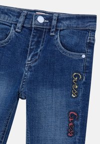 Jeans in denim blu scuro con chiusura a bottone bianco, cinque tasche e loghi "Guess" impreziositi con paillettes dorate e rosse sul davanti.