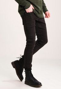 Jeans negros ajustados con una textura suave, con diseño clásico de cinco bolsillos. Combinados con botas de combate negras con cordones. Parte superior verde parcialmente visible.