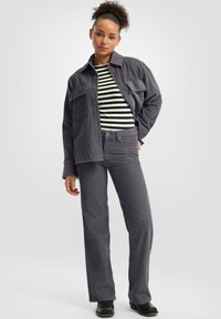 Woman with curly hair wearing a gray corduroy jacket and pants, striped black and white shirt, and black boots, standing against a white background.