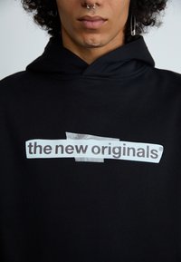 Moleton preto com um gráfico frontal que apresenta o texto "os novos originais" numa etiqueta branca. O material parece suave com uma textura lisa.