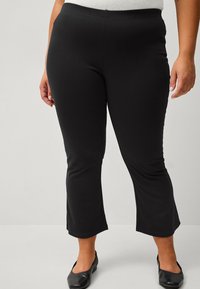 Leggings neri elasticizzati con vita aderente e orlo svasato. Tessuto liscio, senza motivi visibili, abbinati a ballerine nere.