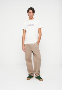 Młody mężczyzna stoi z jedną ręką w kieszeni, ubrany w białą koszulkę Tommy Jeans, beżowe spodnie i zielone sneakersy z brązowymi podeszwami.