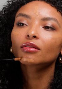 Femme aux cheveux bouclés noirs appliquant du fond de teint sur le menton, le front et les joues à l'aide d'un pinceau de maquillage.
