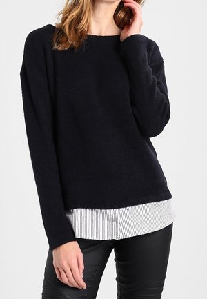 Maglione lavorato a maglia blu navy con collo rotondo e maniche lunghe, indossato sopra una camicia a righe bianche e blu navy. Pantaloni in pelle nera completano l'outfit.