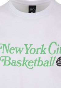 Hvid bomulds t-shirt med grøn tekst, der siger "New York City Basketball", og et sort rundt logo med "kx." Rund halsudskæring.