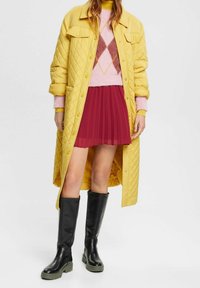 Quilted gul jakke, lyserød argyle sweater, plisseret rød skirt og sorte knæhøje støvler med grønne såler. Minimal hardware, ingen synlige mønstre.