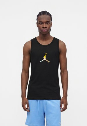 Fiatal férfi fonott hajjal, fekete ujjatlan pólót viselve, amelyen színes Jumpman logó van, és világoskék rövidnadrágban, egyszínű háttér előtt.