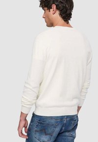 Maglione bianco con maniche lunghe, polsini e orlo a coste. Tessuto morbido, collo tondo, indossato con jeans blu caratterizzati da un design della tasca curva.