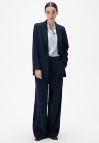 Blazer marine et pantalon large en tissu lisse, associés à une chemise à boutons bleu clair, créant un look ajusté.