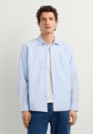 Junger Mann mit lockigem dunklem Haar, der ein hellblaues Hemd über einem weißen T-Shirt und blauen Jeans trägt und vor einem einfarbigen Hintergrund steht.