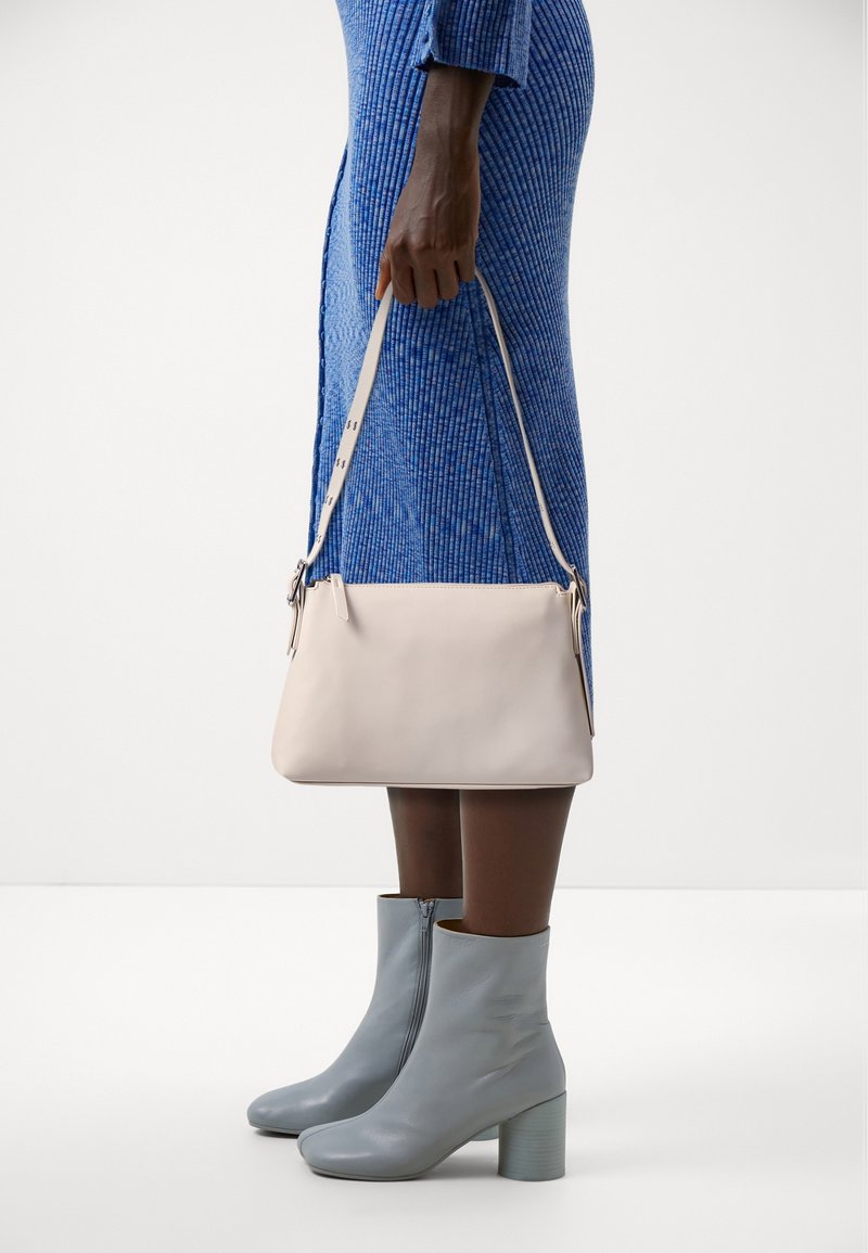 Personne en robe bleue côtelée tenant un sac à main crème, portant des bottines gris clair à talon bloc sur fond blanc.