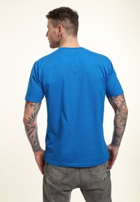 Camiseta de algodón azul con mangas cortas, cuello redondo estándar y una textura suave, que lleva puesta una persona con tatuajes en los brazos.