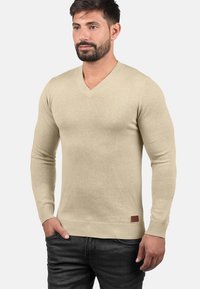 Beżowy sweter v-neck wykonany z dzianiny, z ribbowanymi mankietami i dołem, z małą skórzaną naszywką na dole po lewej stronie.