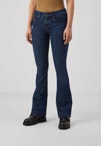 Razširjene temno modre denim hlače z gladko teksturo, zasnovo s petimi žepi, rumenim šivom in kovinskimi gumbi.