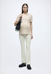 Haut beige texturé à manches courtes, pantalon crème ajusté et chaussures noires épaisses. Le mannequin porte un sac noir avec un détail anneau argenté.