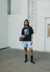 Svart grafisk t-shirt med blå tryck, ljusblå shorts, svarta cowboyboots och en grå crossbody-väska, i en industriell miljö.
